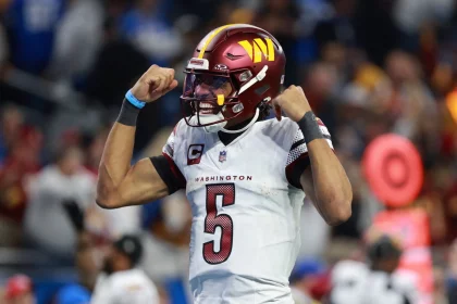 Rookie-Quarterback Jayden Daniels, trägt eine Uniform der Washington Commanders mit der Nummer 5 und feiert. Er trägt ein weißes Trikot, einen burgunderfarbenen Helm und spannt beide Arme an. Der Hintergrund ist verschwommen, einige Personen sind zu sehen und eine rote Seitenlinienmarkierung ist zu sehen. Diese Beschreibung wurde mit der FootballR KI automatisch generiert.