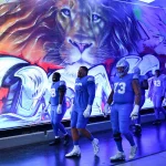 Footballspieler in blauen Uniformen laufen durch einen Tunnel mit einem leuchtenden Löwen-Graffiti, das an die Energie erinnert, die Offensivkoordinator John Morton mitbrachte. Die Helme und Handschuhe der Spieler glänzen unter den Deckenlampen, und ihre nummerierten Trikots tragen zur dynamischen und intensiven Atmosphäre bei. Diese Beschreibung wurde mit der FootballR KI automatisch generiert.