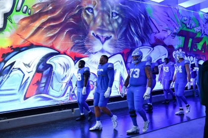 Footballspieler in blauen Uniformen laufen durch einen Tunnel mit einem leuchtenden Löwen-Graffiti, das an die Energie erinnert, die Offensivkoordinator John Morton mitbrachte. Die Helme und Handschuhe der Spieler glänzen unter den Deckenlampen, und ihre nummerierten Trikots tragen zur dynamischen und intensiven Atmosphäre bei. Diese Beschreibung wurde mit der FootballR KI automatisch generiert.