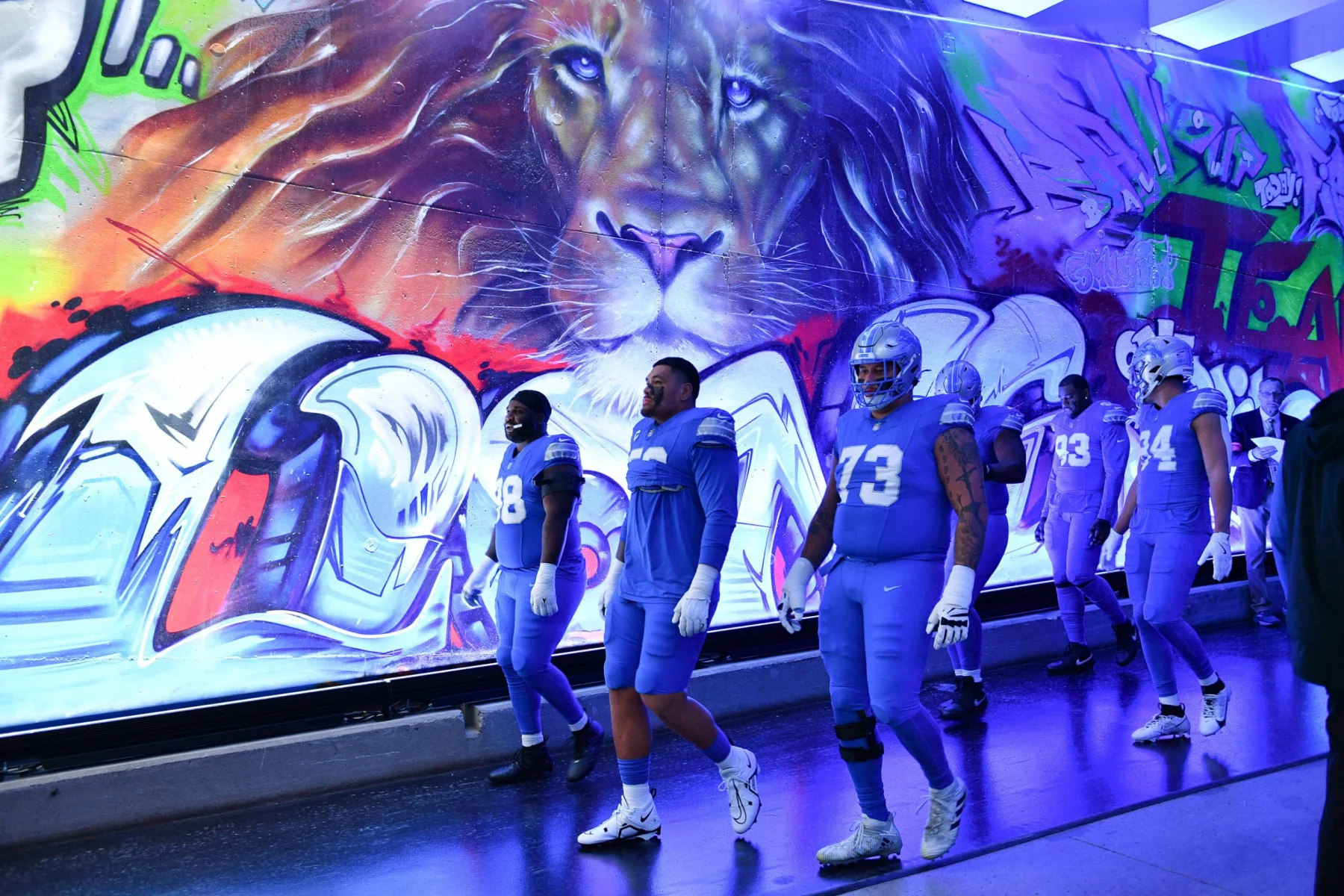 Footballspieler in blauen Uniformen laufen durch einen Tunnel mit einem leuchtenden Löwen-Graffiti, das an die Energie erinnert, die Offensivkoordinator John Morton mitbrachte. Die Helme und Handschuhe der Spieler glänzen unter den Deckenlampen, und ihre nummerierten Trikots tragen zur dynamischen und intensiven Atmosphäre bei. Diese Beschreibung wurde mit der FootballR KI automatisch generiert.