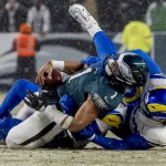 Im Meisterschaftsspiel, bei dem alles auf dem Spiel steht, wird Jalen Hurts in blaugrüner und schwarzer Uniform auf dem schneebedeckten Feld von zwei gegnerischen Spielern in blau-weißen Uniformen angegriffen. Der angegriffene Spieler hält den Ball fest, während Schneeflocken umherwirbeln, während im Hintergrund das Stadionpersonal unscharf zu sehen ist. Diese Beschreibung wurde mit der FootballR KI automatisch generiert.
