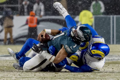 Im Meisterschaftsspiel, bei dem alles auf dem Spiel steht, wird Jalen Hurts in blaugrüner und schwarzer Uniform auf dem schneebedeckten Feld von zwei gegnerischen Spielern in blau-weißen Uniformen angegriffen. Der angegriffene Spieler hält den Ball fest, während Schneeflocken umherwirbeln, während im Hintergrund das Stadionpersonal unscharf zu sehen ist. Diese Beschreibung wurde mit der FootballR KI automatisch generiert.