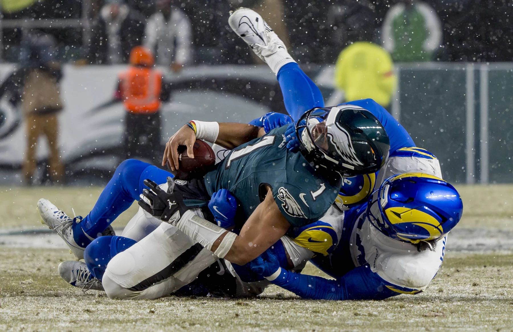 Im Meisterschaftsspiel, bei dem alles auf dem Spiel steht, wird Jalen Hurts in blaugrüner und schwarzer Uniform auf dem schneebedeckten Feld von zwei gegnerischen Spielern in blau-weißen Uniformen angegriffen. Der angegriffene Spieler hält den Ball fest, während Schneeflocken umherwirbeln, während im Hintergrund das Stadionpersonal unscharf zu sehen ist. Diese Beschreibung wurde mit der FootballR KI automatisch generiert.