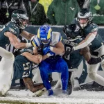 Ein Footballspieler in Blau und Gelb wird auf einem schneebedeckten Feld von vier Philadelphia Eagles Spielern in grün-weißen Trikots angegriffen. Die konzentrierten und intensiven Athleten liefern sich einen erbitterten Kampf unter wirbelnden Schneeflocken, was die kalten, winterlichen Bedingungen dieses Divisional-Round-Spiels unterstreicht. Diese Beschreibung wurde mit der FootballR KI automatisch generiert.