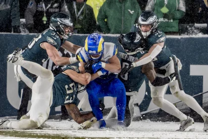 Ein Footballspieler in Blau und Gelb wird auf einem schneebedeckten Feld von vier Philadelphia Eagles Spielern in grün-weißen Trikots angegriffen. Die konzentrierten und intensiven Athleten liefern sich einen erbitterten Kampf unter wirbelnden Schneeflocken, was die kalten, winterlichen Bedingungen dieses Divisional-Round-Spiels unterstreicht. Diese Beschreibung wurde mit der FootballR KI automatisch generiert.