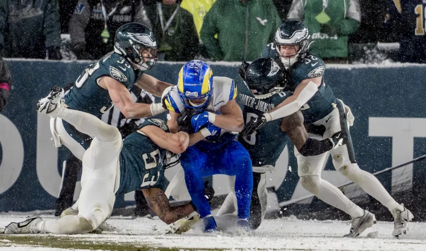 Ein Footballspieler in Blau und Gelb wird auf einem schneebedeckten Feld von vier Philadelphia Eagles Spielern in grün-weißen Trikots angegriffen. Die konzentrierten und intensiven Athleten liefern sich einen erbitterten Kampf unter wirbelnden Schneeflocken, was die kalten, winterlichen Bedingungen dieses Divisional-Round-Spiels unterstreicht. Diese Beschreibung wurde mit der FootballR KI automatisch generiert.
