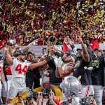 Eine siegreiche Footballmannschaft feiert auf dem Feld und sonnt sich im Ruhm des National Championship Game. Die Spieler der Ohio State Buckeyes tragen rot-weiße Trikots mit schwarzen Kappen und goldenem Konfetti. Im Hintergrund jubeln die Fans wild, während Fotografen den ikonischen Moment eifrig einfangen. Diese Beschreibung wurde mit der FootballR KI automatisch generiert.