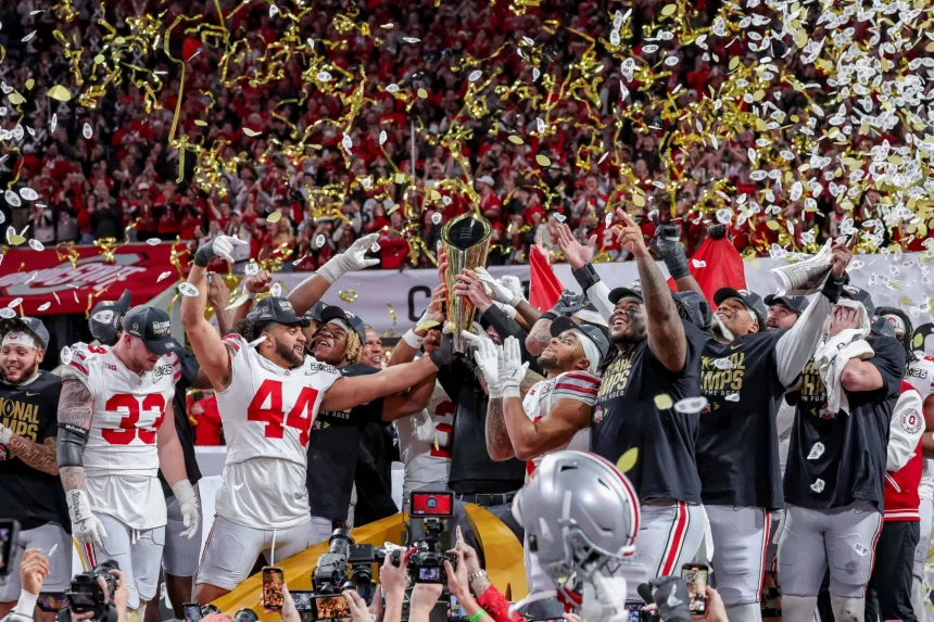 Eine siegreiche Footballmannschaft feiert auf dem Feld und sonnt sich im Ruhm des National Championship Game. Die Spieler der Ohio State Buckeyes tragen rot-weiße Trikots mit schwarzen Kappen und goldenem Konfetti. Im Hintergrund jubeln die Fans wild, während Fotografen den ikonischen Moment eifrig einfangen. Diese Beschreibung wurde mit der FootballR KI automatisch generiert.