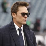 Tom Brady mit Sonnenbrille und kariertem Mantel steht im Freien vor verschwommenen Stadionsitzen. Er hat braunes Haar und trägt eine dunkle Krawatte über einer Weste. Angesichts seiner Kleidung und des bewölkten Himmels wirkt die Atmosphäre kühl. Diese Beschreibung wurde mit der FootballR KI automatisch generiert.