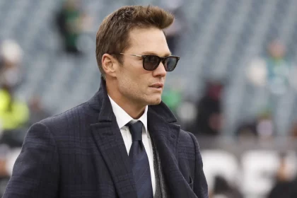Tom Brady mit Sonnenbrille und kariertem Mantel steht im Freien vor verschwommenen Stadionsitzen. Er hat braunes Haar und trägt eine dunkle Krawatte über einer Weste. Angesichts seiner Kleidung und des bewölkten Himmels wirkt die Atmosphäre kühl. Diese Beschreibung wurde mit der FootballR KI automatisch generiert.