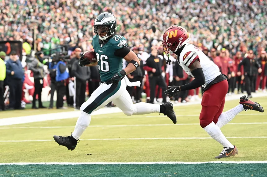 Ein Footballspieler im Trikot der Philadelphia Eagles, Saquon Barkley (Nummer 26), rennt während des Super Bowl LIX in Richtung Endzone. Ein rot-weiß uniformierter Gegner kämpft darum, ihn einzuholen, während die Menge im Stadion zu einer verschwommenen Kulisse für diesen spannenden Moment wird. Diese Beschreibung wurde mit der FootballR KI automatisch generiert.