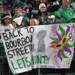 Eine Gruppe von Philadelphia Eagles Fans in festlicher Kleidung hält ein handgemaltes Banner mit der Aufschrift „Zurück zur Bourbon Street!! Lasst uns jagen!!“. Auf dem Banner sind ein Footballpokal und farbenfrohe Mardi Gras-Kunstwerke abgebildet. Die Menge ist in den Teamfarben gekleidet, einige tragen Gesichtsbemalung oder Kostüme und verkörpern damit den Geist von New Orleans. Diese Beschreibung wurde mit der FootballR KI automatisch generiert.