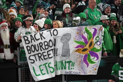 Eine Gruppe von Philadelphia Eagles Fans in festlicher Kleidung hält ein handgemaltes Banner mit der Aufschrift „Zurück zur Bourbon Street!! Lasst uns jagen!!“. Auf dem Banner sind ein Footballpokal und farbenfrohe Mardi Gras-Kunstwerke abgebildet. Die Menge ist in den Teamfarben gekleidet, einige tragen Gesichtsbemalung oder Kostüme und verkörpern damit den Geist von New Orleans. Diese Beschreibung wurde mit der FootballR KI automatisch generiert.