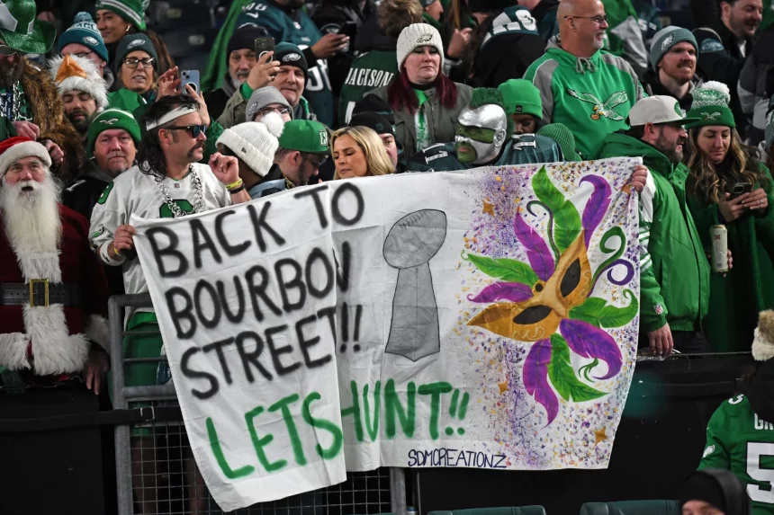 Eine Gruppe von Philadelphia Eagles Fans in festlicher Kleidung hält ein handgemaltes Banner mit der Aufschrift „Zurück zur Bourbon Street!! Lasst uns jagen!!“. Auf dem Banner sind ein Footballpokal und farbenfrohe Mardi Gras-Kunstwerke abgebildet. Die Menge ist in den Teamfarben gekleidet, einige tragen Gesichtsbemalung oder Kostüme und verkörpern damit den Geist von New Orleans. Diese Beschreibung wurde mit der FootballR KI automatisch generiert.