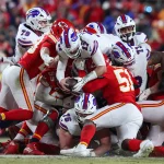 Heute im TV - Footballspieler zweier Teams, eines in Rot und das andere in Weiß mit blauen und roten Akzenten, die an die Buffalo Bills erinnern, stehen an der Line of Scrimmage in einem Haufen. Ein Spieler in Weiß hält den Ball, während er angegriffen wird. Im Hintergrund ist eine verschwommene Menschenmenge zu sehen, was die intensive Action der Szene noch verstärkt. Diese Beschreibung wurde mit der FootballR KI automatisch generiert.