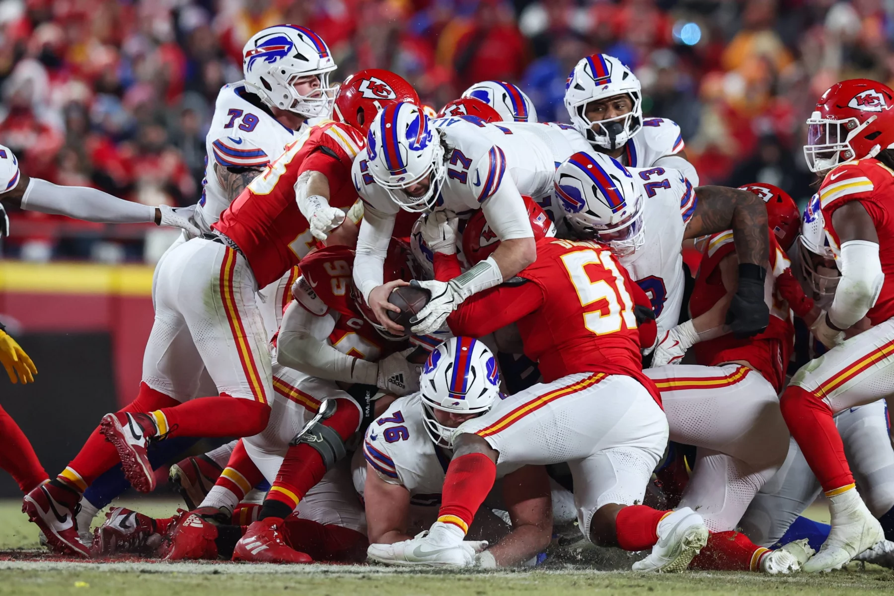 Heute im TV - Footballspieler zweier Teams, eines in Rot und das andere in Weiß mit blauen und roten Akzenten, die an die Buffalo Bills erinnern, stehen an der Line of Scrimmage in einem Haufen. Ein Spieler in Weiß hält den Ball, während er angegriffen wird. Im Hintergrund ist eine verschwommene Menschenmenge zu sehen, was die intensive Action der Szene noch verstärkt. Diese Beschreibung wurde mit der FootballR KI automatisch generiert.