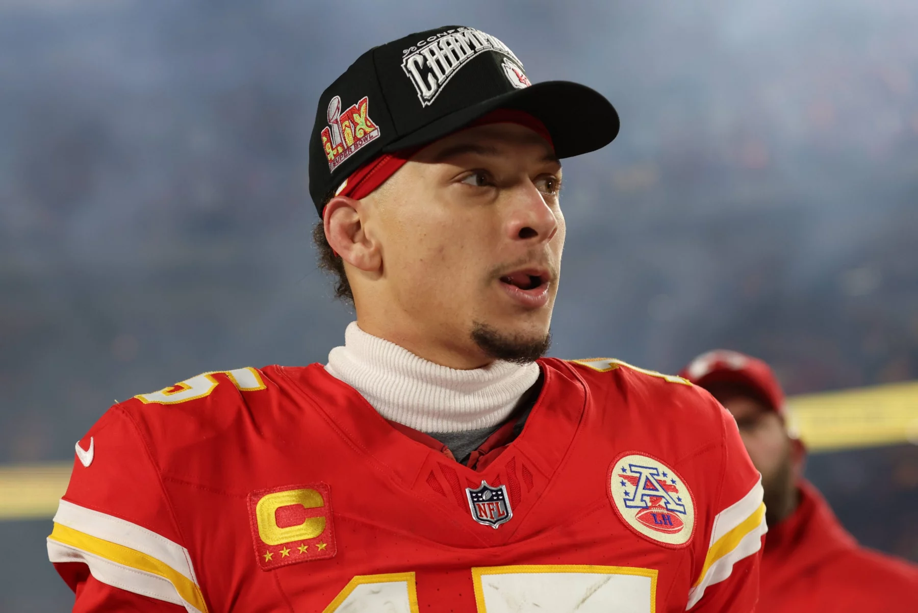 Footballspieler Patrick Mahomes steht in der roten Uniform der Kansas City Chiefs auf dem Feld und trägt eine schwarze Mütze mit dem Schriftzug „Super Bowl Champions“. Er trägt einen weißen Rollkragenpullover und ein rotes Trikot mit der Nummer 15, umgeben von verschwommenen Stadionlichtern und jubelnden Zuschauern im Hintergrund. Diese Beschreibung wurde mit der FootballR KI automatisch generiert.