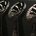 Super Bowl LIX: Drei glänzende Vince Lombardi-Trophäen glänzen unter den hellen Lichtern und werden vor einem schwarzen Hintergrund präsentiert. Die reflektierenden silbernen Oberflächen zeigen ein Muster aus leuchtenden Linien und Punkten und feiern den Ruhm des Super Bowl LIX, wobei auf jedem hohen Sockel majestätisch ein Football thront. Diese Beschreibung wurde mit der FootballR KI automatisch generiert.
