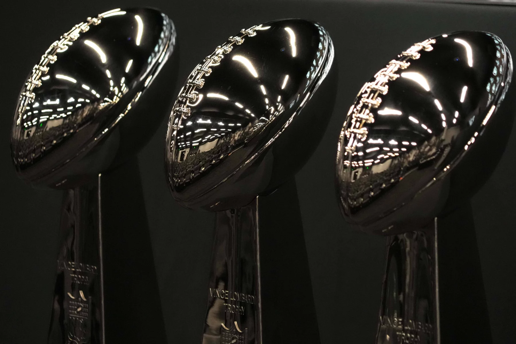 Super Bowl LIX: Drei glänzende Vince Lombardi-Trophäen glänzen unter den hellen Lichtern und werden vor einem schwarzen Hintergrund präsentiert. Die reflektierenden silbernen Oberflächen zeigen ein Muster aus leuchtenden Linien und Punkten und feiern den Ruhm des Super Bowl LIX, wobei auf jedem hohen Sockel majestätisch ein Football thront. Diese Beschreibung wurde mit der FootballR KI automatisch generiert.