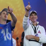 Zwei Männer stehen im Freien und zeigen nach oben. Der Mann links trägt ein blaues Trikot mit der Nummer 16, Jared Goff. Eli Manning rechts trägt einen weißen Pullover und ein Namensschild. Lebendige abstrakte Designs füllen den Hintergrund, während andere in der Ferne zu sehen sind. Diese Beschreibung wurde mit der FootballR KI automatisch generiert.