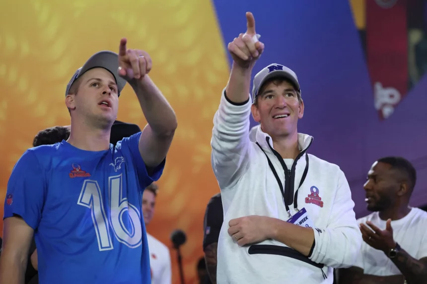 Zwei Männer stehen im Freien und zeigen nach oben. Der Mann links trägt ein blaues Trikot mit der Nummer 16, Jared Goff. Eli Manning rechts trägt einen weißen Pullover und ein Namensschild. Lebendige abstrakte Designs füllen den Hintergrund, während andere in der Ferne zu sehen sind. Diese Beschreibung wurde mit der FootballR KI automatisch generiert.