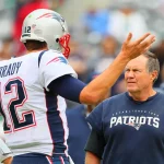 Tom Brady in der Uniform der New England Patriots, Nummer 12, gestikuliert, während er sich mit Trainer Bill Belichick im Patriots-Sweatshirt berät. Sie stehen auf dem Spielfeld, hinter ihnen sind verschwommene Zuschauer, die wahrscheinlich Strategien für das Spiel entwickeln. Diese Beschreibung wurde mit der FootballR KI automatisch generiert.