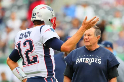 Tom Brady in der Uniform der New England Patriots, Nummer 12, gestikuliert, während er sich mit Trainer Bill Belichick im Patriots-Sweatshirt berät. Sie stehen auf dem Spielfeld, hinter ihnen sind verschwommene Zuschauer, die wahrscheinlich Strategien für das Spiel entwickeln. Diese Beschreibung wurde mit der FootballR KI automatisch generiert.
