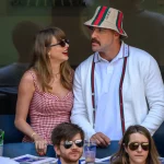 Taylor Swift in einem rotkarierten Kleid und Sonnenbrille strahlt Travis Kelce in weißer Strickjacke, Anglerhut und Schnurrbart an. Diese Beschreibung wurde mit der FootballR KI automatisch generiert.