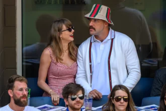 Travis Kelce - Taylor Swift in einem rotkarierten Kleid und Sonnenbrille strahlt Travis Kelce in weißer Strickjacke, Anglerhut und Schnurrbart an. Diese Beschreibung wurde mit der FootballR KI automatisch generiert.