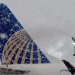 Kansas City Chiefs: Ein Arbeiter sprüht bei starkem Schneefall Enteisungsmittel auf das Heck eines Flugzeugs. Das Heck, das ein blaues Globus-Design aufweist, das an den mutigen Geist der Kansas City Chiefs erinnert, ist von großen Schneeflocken umhüllt und sorgt so für eine winterliche Szenerie auf dem geschäftigen Flughafen. Diese Beschreibung wurde mit der FootballR KI automatisch generiert.