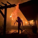Silhouette eines Feuerwehrmanns mit Schlauch in der Hand, der auf einen großen Brand zugeht, der nachts ein Haus in Los Angeles verschlingt. Die Szene wird von intensiven orangefarbenen Flammen beleuchtet, mit dunklen Schatten einer hölzernen Pergola im Vordergrund. Der Himmel ist voller Rauch, der an die jüngsten Waldbrände erinnert und die Dringlichkeit der Situation unterstreicht. Diese Beschreibung wurde mit der FootballR KI automatisch generiert.