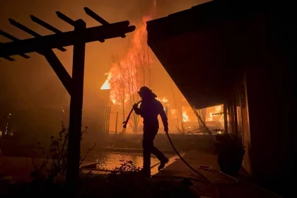 Silhouette eines Feuerwehrmanns mit Schlauch in der Hand, der auf einen großen Brand zugeht, der nachts ein Haus in Los Angeles verschlingt. Die Szene wird von intensiven orangefarbenen Flammen beleuchtet, mit dunklen Schatten einer hölzernen Pergola im Vordergrund. Der Himmel ist voller Rauch, der an die jüngsten Waldbrände erinnert und die Dringlichkeit der Situation unterstreicht. Diese Beschreibung wurde mit der FootballR KI automatisch generiert.