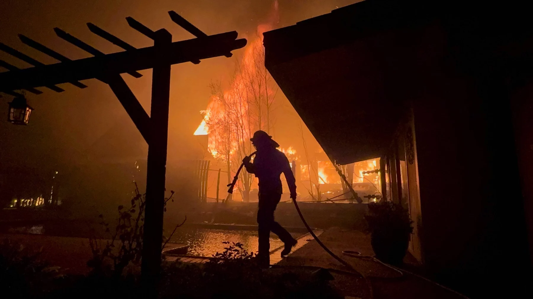 Silhouette eines Feuerwehrmanns mit Schlauch in der Hand, der auf einen großen Brand zugeht, der nachts ein Haus in Los Angeles verschlingt. Die Szene wird von intensiven orangefarbenen Flammen beleuchtet, mit dunklen Schatten einer hölzernen Pergola im Vordergrund. Der Himmel ist voller Rauch, der an die jüngsten Waldbrände erinnert und die Dringlichkeit der Situation unterstreicht. Diese Beschreibung wurde mit der FootballR KI automatisch generiert.