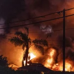 Wild Card Round: Ein nächtliches Bild eines großen Brandes, ein echter Überraschungseffekt, verschlingt ein Wohngebiet. Hohe Flammen und dichter Rauch erheben sich gegen den dunklen Himmel, während die Silhouette einer Palme davor steht. Straßenschilder weisen auf eine Geschwindigkeitsbegrenzung von 30 hin, im Vordergrund sind Radarüberwachungssysteme zu sehen. Diese Beschreibung wurde mit der FootballR KI automatisch generiert.