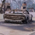 Ein verkohltes Auto steht auf einer Einfahrt in Los Angeles, zerstört durch einen Brand. Die umliegenden Bäume sind verbrannt und haben kahle Äste. Im Hintergrund sind ein gemauerter Schornstein und beschädigte Gebäude zu sehen, vor einem durch Rauch verhangenen Himmel. Im Vordergrund ist ein rissiger Bürgersteig mit verstreuten Trümmern zu sehen. Diese Beschreibung wurde mit der FootballR KI automatisch generiert.