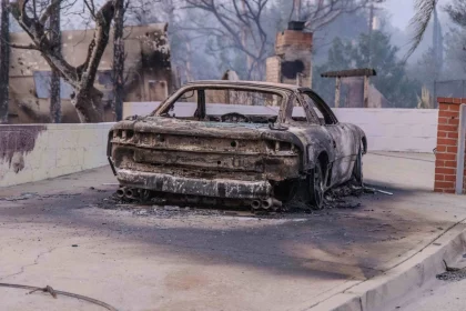 Ein verkohltes Auto steht auf einer Einfahrt in Los Angeles, zerstört durch einen Brand. Die umliegenden Bäume sind verbrannt und haben kahle Äste. Im Hintergrund sind ein gemauerter Schornstein und beschädigte Gebäude zu sehen, vor einem durch Rauch verhangenen Himmel. Im Vordergrund ist ein rissiger Bürgersteig mit verstreuten Trümmern zu sehen. Diese Beschreibung wurde mit der FootballR KI automatisch generiert.