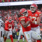 Zwei Spieler der Kansas City Chiefs in roten Uniformen feiern einen Spielzug auf dem Spielfeld. Der Spieler links hält den Ball, während der andere ihn umarmt. Eine verschwommene Menschenmenge in den Farben der Chiefs füllt den Hintergrund. Zu sehen sind Spieler der gegnerischen Mannschaft und eine Stadionwerbung. Diese Beschreibung wurde mit der FootballR KI automatisch generiert.