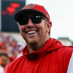 Liam Coen mit roter Tampa Bay Buccaneers-Kappe und Sonnenbrille lächelt und trägt eine rote ärmellose Jacke über einem weißen Langarmshirt. Im Hintergrund ist ein verschwommenes Stadion voller Zuschauer und großer Bildschirme zu sehen. Könnte das Liam Coen sein, der den Spieltag genießt?. Diese Beschreibung wurde mit der FootballR KI automatisch generiert.