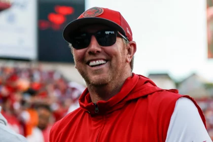 Liam Coen mit roter Tampa Bay Buccaneers-Kappe und Sonnenbrille lächelt und trägt eine rote ärmellose Jacke über einem weißen Langarmshirt. Im Hintergrund ist ein verschwommenes Stadion voller Zuschauer und großer Bildschirme zu sehen. Könnte das Liam Coen sein, der den Spieltag genießt?. Diese Beschreibung wurde mit der FootballR KI automatisch generiert.