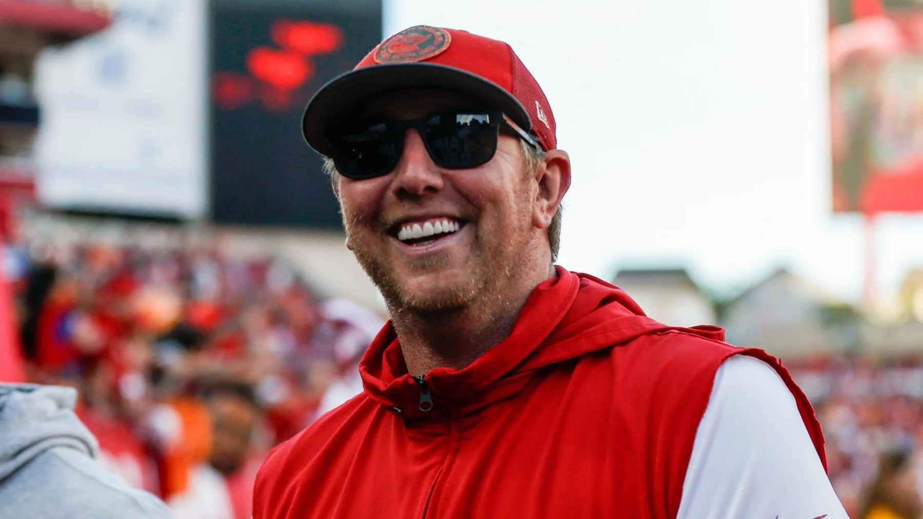 Liam Coen mit roter Tampa Bay Buccaneers-Kappe und Sonnenbrille lächelt und trägt eine rote ärmellose Jacke über einem weißen Langarmshirt. Im Hintergrund ist ein verschwommenes Stadion voller Zuschauer und großer Bildschirme zu sehen. Könnte das Liam Coen sein, der den Spieltag genießt?. Diese Beschreibung wurde mit der FootballR KI automatisch generiert.