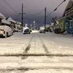 Eine schneebedeckte Wohnstraße bei Nacht erinnert an einen ruhigen Abend in New Orleans während des seltenen Schneesturms. Autos stehen geparkt, teilweise unter Schnee begraben, während sich Stromleitungen unter einem bedeckten Himmel durch die Szene ziehen. Spuren sind sichtbar und markieren, wo das Leben inmitten des ruhigen Schneefalls weitergeht. Diese Beschreibung wurde mit der FootballR KI automatisch generiert.