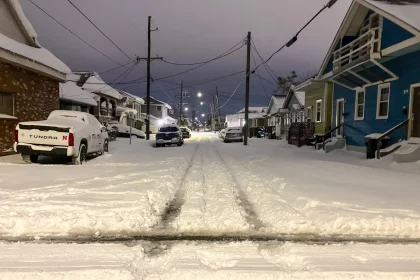 Eine schneebedeckte Wohnstraße bei Nacht erinnert an einen ruhigen Abend in New Orleans während des seltenen Schneesturms. Autos stehen geparkt, teilweise unter Schnee begraben, während sich Stromleitungen unter einem bedeckten Himmel durch die Szene ziehen. Spuren sind sichtbar und markieren, wo das Leben inmitten des ruhigen Schneefalls weitergeht. Diese Beschreibung wurde mit der FootballR KI automatisch generiert.