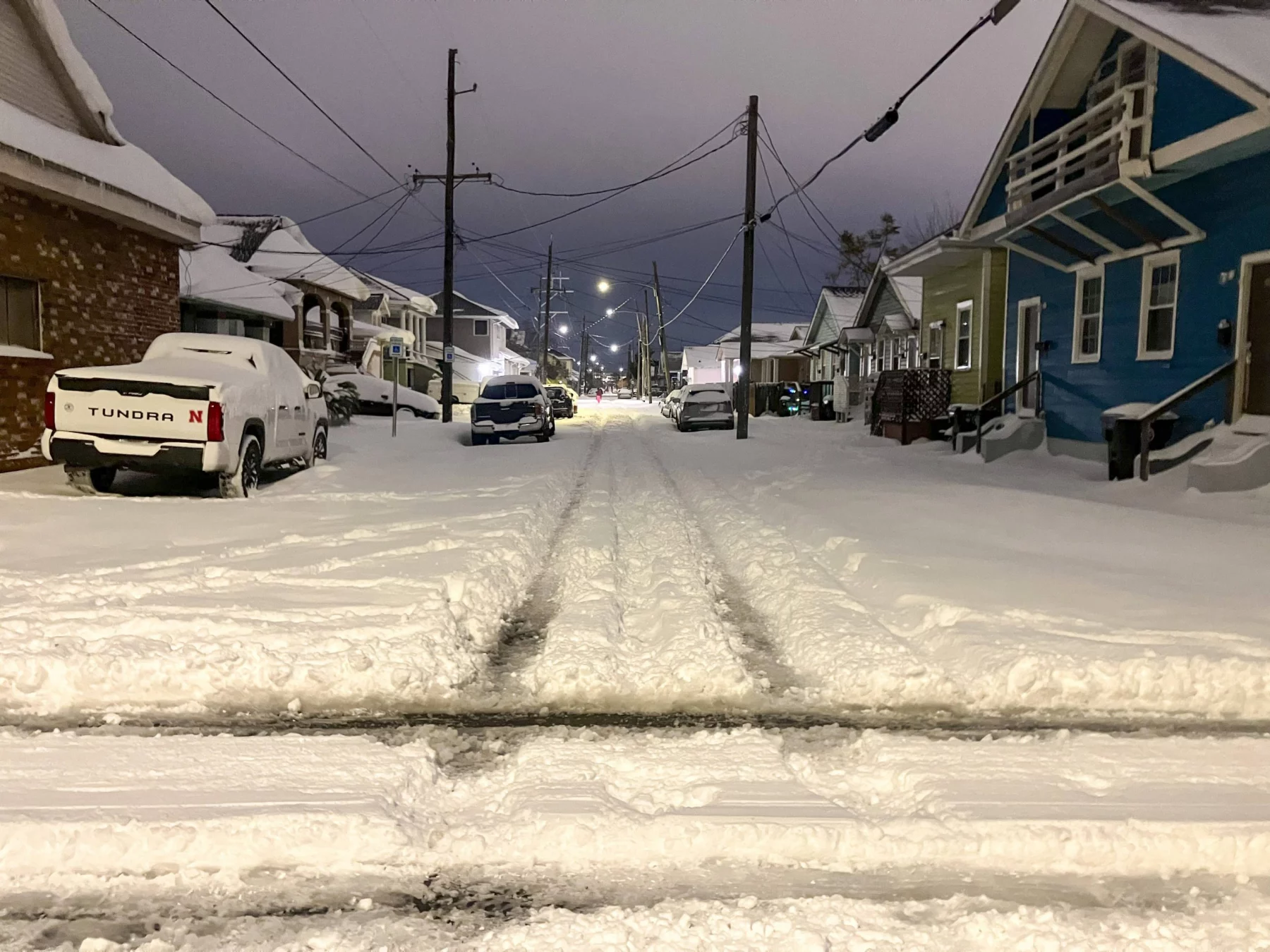 Eine schneebedeckte Wohnstraße bei Nacht erinnert an einen ruhigen Abend in New Orleans während des seltenen Schneesturms. Autos stehen geparkt, teilweise unter Schnee begraben, während sich Stromleitungen unter einem bedeckten Himmel durch die Szene ziehen. Spuren sind sichtbar und markieren, wo das Leben inmitten des ruhigen Schneefalls weitergeht. Diese Beschreibung wurde mit der FootballR KI automatisch generiert.