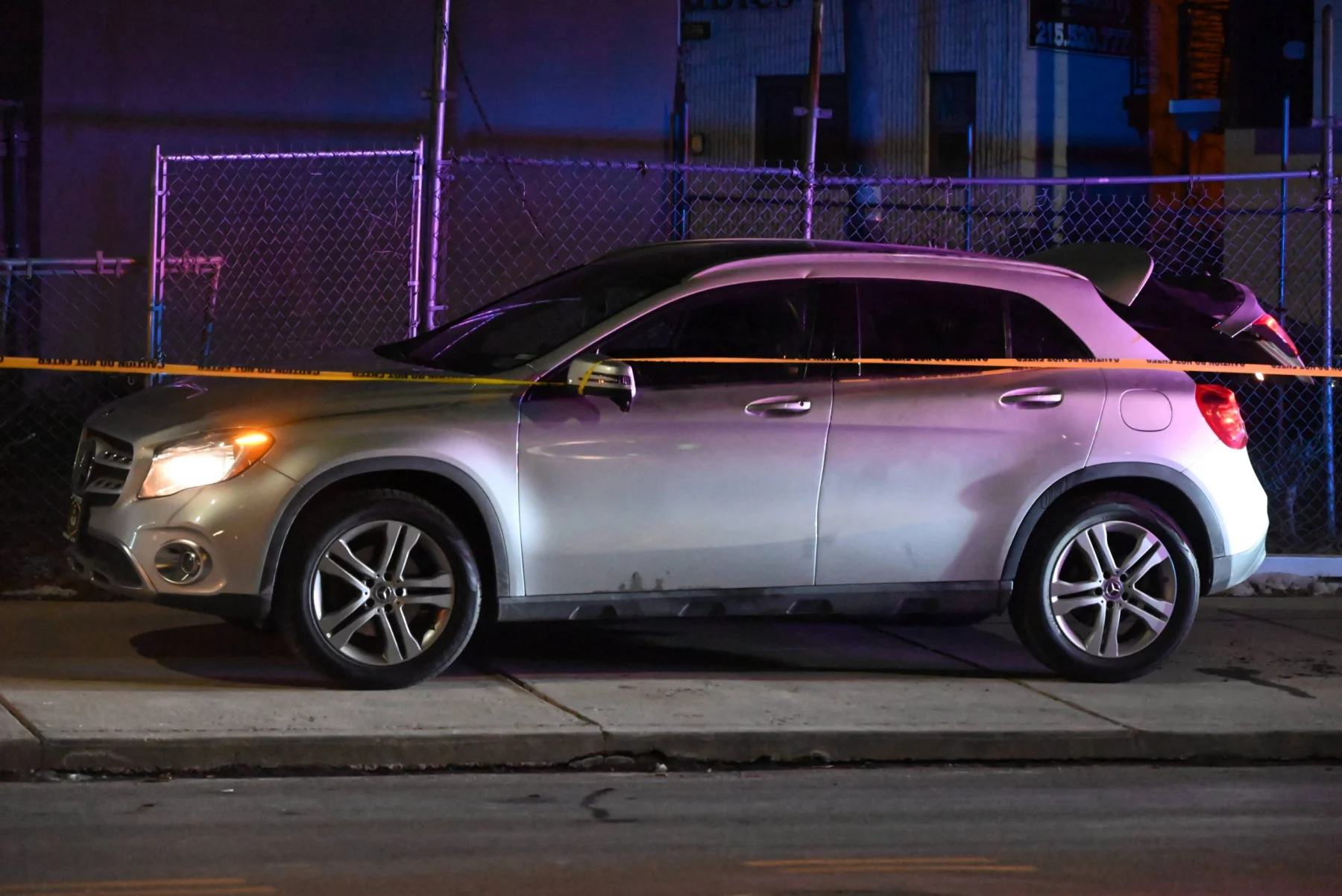Nach dem Championship Game: Ein silberner SUV steht nachts auf einer schwach beleuchteten Straße, die an ein spannendes Meisterschaftsspiel erinnert. Polizeiabsperrungen spannen sich über die gesamte Länge und kennzeichnen den Tatort. Im Hintergrund spiegeln sich bunte Lichter eines nahe gelegenen Gebäudes in der Oberfläche des Autos und werfen einen unheimlichen Schein durch den Maschendrahtzaun. Diese Beschreibung wurde mit der FootballR KI automatisch generiert.