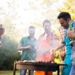 NFL Accessoires - Eine Gruppe von sechs Freunden genießt eine lebhafte Grillparty im Freien. Zwei tragen Schürzen und kochen auf einem Grill, um sie herum steigt Rauch auf. Die anderen lächeln und unterhalten sich in ihrer lässigen Sommerkleidung vor einer grünen, blattreichen Kulisse, die an eine Park- oder Gartenkulisse erinnert. Diese Beschreibung wurde mit der FootballR KI automatisch generiert.