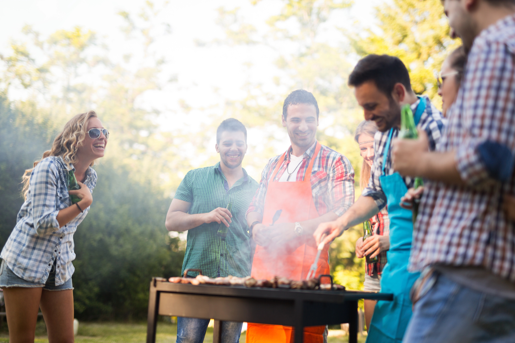 NFL Accessoires - Eine Gruppe von sechs Freunden genießt eine lebhafte Grillparty im Freien. Zwei tragen Schürzen und kochen auf einem Grill, um sie herum steigt Rauch auf. Die anderen lächeln und unterhalten sich in ihrer lässigen Sommerkleidung vor einer grünen, blattreichen Kulisse, die an eine Park- oder Gartenkulisse erinnert. Diese Beschreibung wurde mit der FootballR KI automatisch generiert.