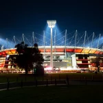 Melbourne Cricket Ground bekommt im Rahmen der International Series ein NFL Spiel. Beleuchtetes Stadion bei Nacht mit hellen Flutlichtern und einer markanten Dachkonstruktion vor einem dunklen Himmel. Das Äußere ist in Rottönen beleuchtet und im Vordergrund sind Bäume zu sehen. In der Nähe des Eingangs sind Silhouetten von Menschen zu sehen, die eine lebendige Atmosphäre schaffen. Diese Beschreibung wurde mit der FootballR KI automatisch generiert.