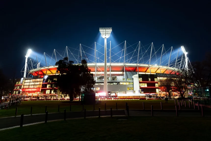 Melbourne Cricket Ground bekommt im Rahmen der International Series ein NFL Spiel. Beleuchtetes Stadion bei Nacht mit hellen Flutlichtern und einer markanten Dachkonstruktion vor einem dunklen Himmel. Das Äußere ist in Rottönen beleuchtet und im Vordergrund sind Bäume zu sehen. In der Nähe des Eingangs sind Silhouetten von Menschen zu sehen, die eine lebendige Atmosphäre schaffen. Diese Beschreibung wurde mit der FootballR KI automatisch generiert.