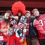 Vier Männer in auffälliger Tampa Bay Buccaneers-Ausrüstung posieren gemeinsam auf dem roten Teppich und strahlen Begeisterung für das NFL International Event aus. Einer trägt eine große rote Afroperücke und einen Anzug mit Augenmotiven, während ein anderer eine bunte Jacke trägt. Zwei tragen stolz rote Trikots und feiern ihren Erfolg beim Wettbewerb „Fan des Jahres“. Diese Beschreibung wurde mit der FootballR KI automatisch generiert.