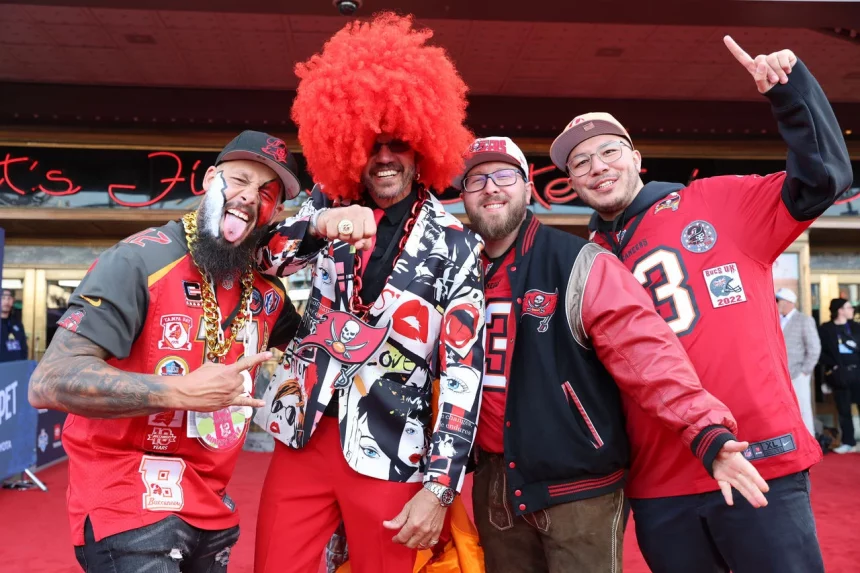 Vier Männer in auffälliger Tampa Bay Buccaneers-Ausrüstung posieren gemeinsam auf dem roten Teppich und strahlen Begeisterung für das NFL International Event aus. Einer trägt eine große rote Afroperücke und einen Anzug mit Augenmotiven, während ein anderer eine bunte Jacke trägt. Zwei tragen stolz rote Trikots und feiern ihren Erfolg beim Wettbewerb „Fan des Jahres“. Diese Beschreibung wurde mit der FootballR KI automatisch generiert.